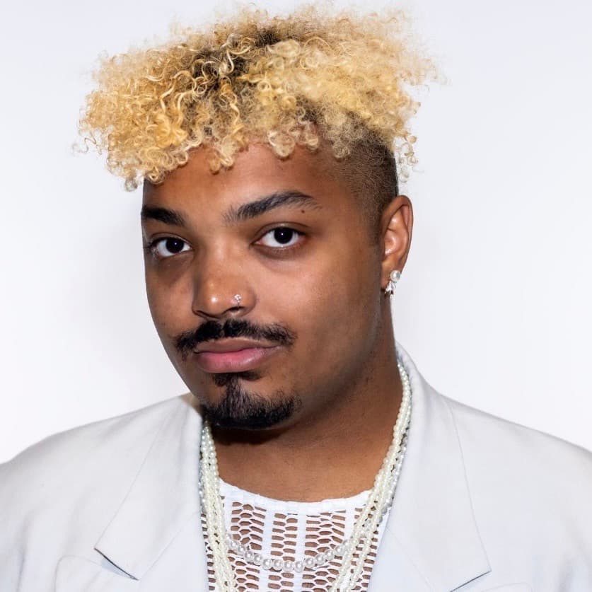 Portrait of Trey May, wearing a mesh top, light blazer, and pearl necklace against a white studio background.